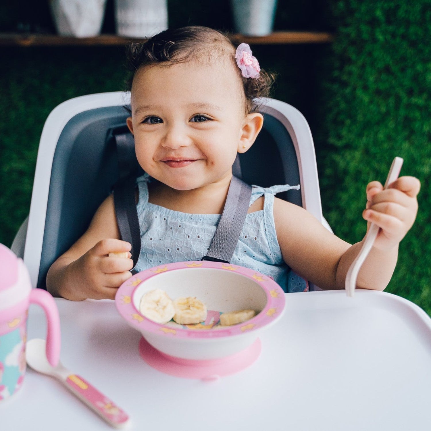 Smiling toddler sitting in a high chair using a pink suction bowl while practicing self-feeding with a spoon. A matching training cup and utensil are placed on the tray, creating a clean and engaging mealtime scene.