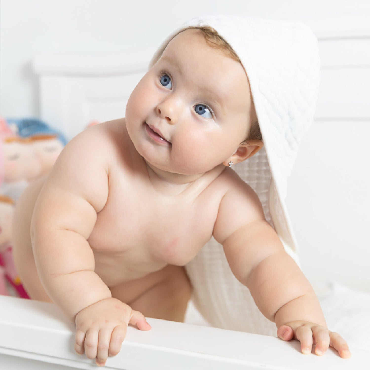 A smiling baby leans over the edge of a bathtub with a soft white hooded muslin towel draped over their head after bath time. The lightweight, ultra-absorbent towel is designed to keep babies warm and dry while being gentle on delicate skin.