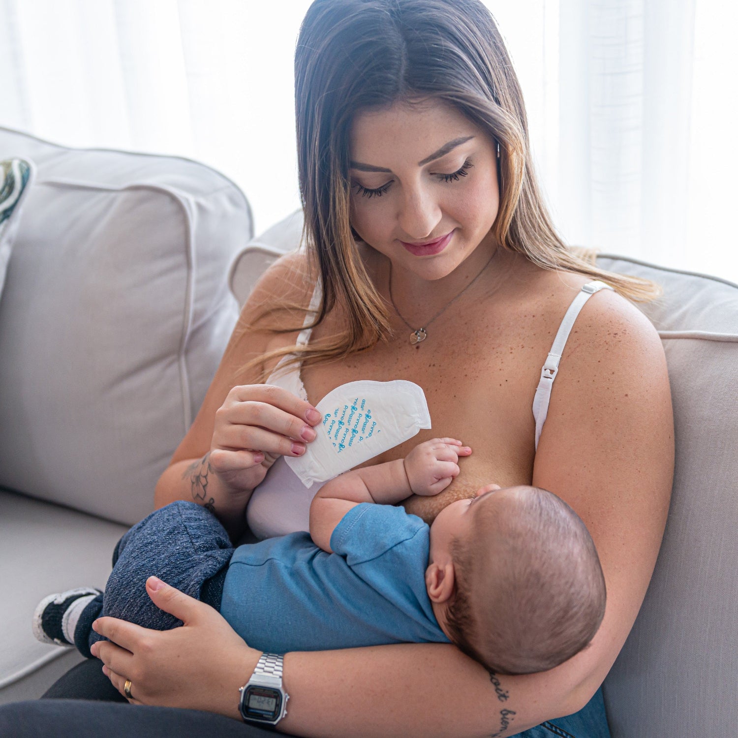 A mother breastfeeding her baby while holding a Primo Passi disposable nursing pad. The ultra-absorbent, contoured nursing pad helps protect against leaks between feedings, providing discreet, breathable comfort for breastfeeding moms throughout the day.