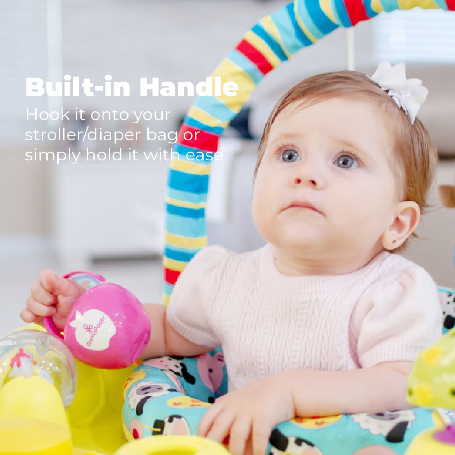 Baby holding a Primo Passi pink pacifier case by its built-in handle while sitting in a play seat. The BPA-free case is designed to attach easily to a stroller, diaper bag, or handbag, keeping pacifiers clean, protected, and within easy reach wherever you go.