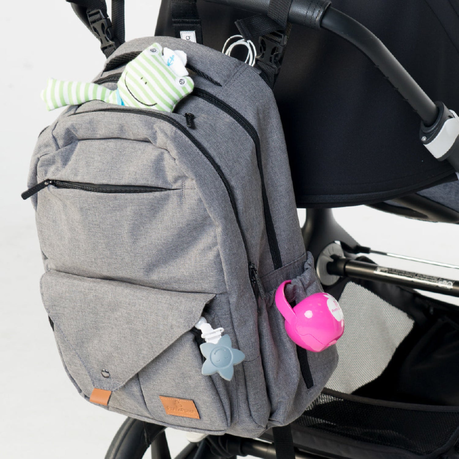 Gray diaper backpack attached to a stroller with a Primo Passi pink pacifier case clipped to the side pocket using its built-in handle. The compact, BPA-free case keeps pacifiers clean, protected, and easily accessible while traveling with baby.