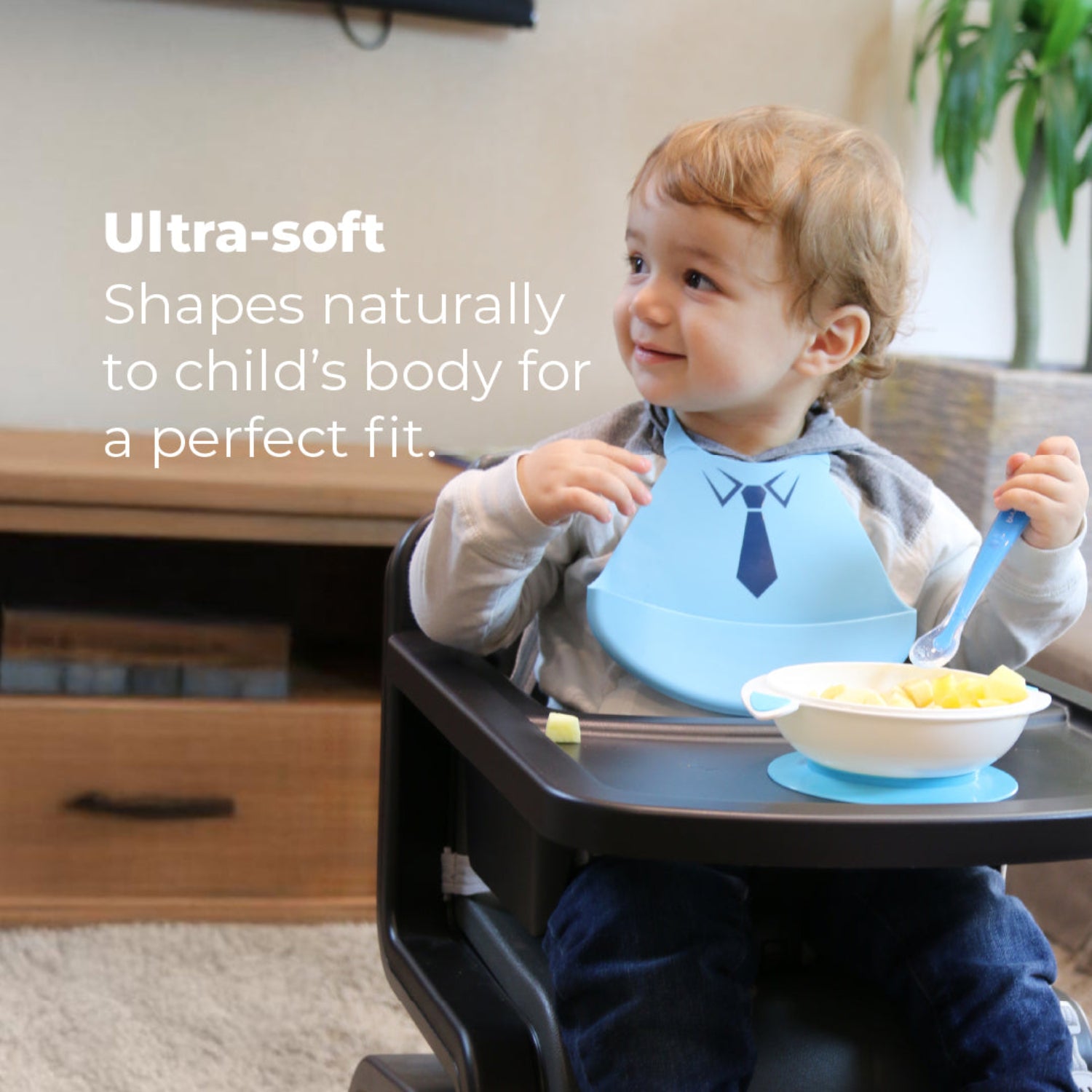 Smiling toddler seated in a high chair wearing a blue Primo Passi silicone bib with a tuxedo design while eating from a white suction bowl filled with fruit. The child holds a blue spoon, and the image highlights the bib's ultra-soft silicone that naturally conforms to the child's body for a comfortable fit during mealtime.