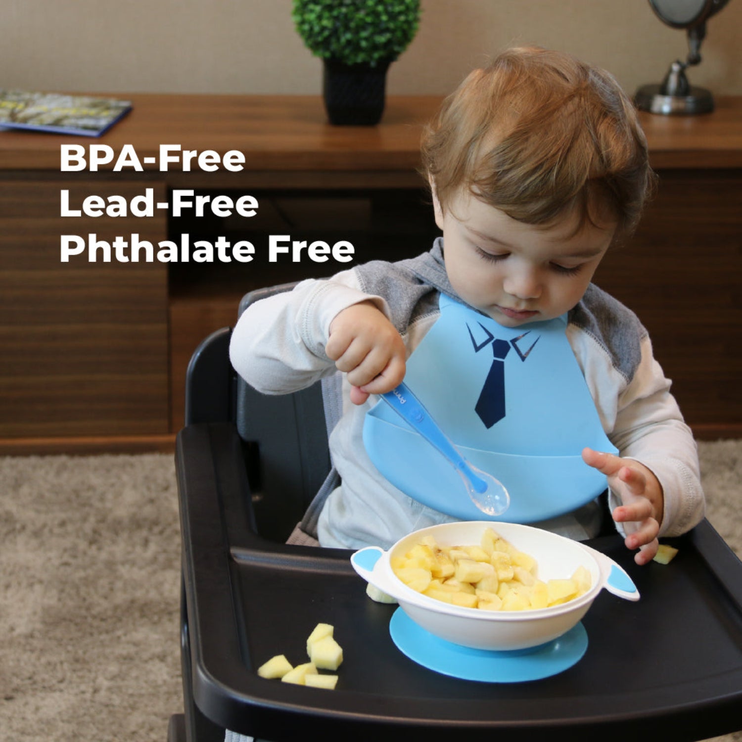 Toddler sitting in a high chair wearing a blue Primo Passi silicone bib with a tuxedo design while eating diced fruit from a white suction bowl. The child holds a blue spoon and practices self-feeding, while the image highlights the bib's BPA-free, lead-free, and phthalate-free materials for safe everyday mealtimes.