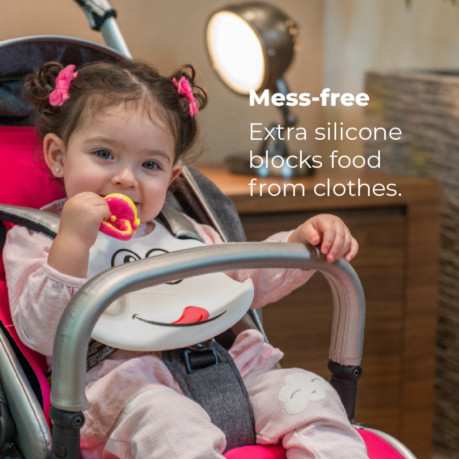 Toddler sitting in a stroller wearing a white Primo Passi silicone bib with a playful face design while holding a colorful teething toy. The deep catch pocket helps keep food off clothing, making mealtime cleaner at home or on the go. The cozy indoor setting highlights the bib's flexible, waterproof silicone material and mess-free design.