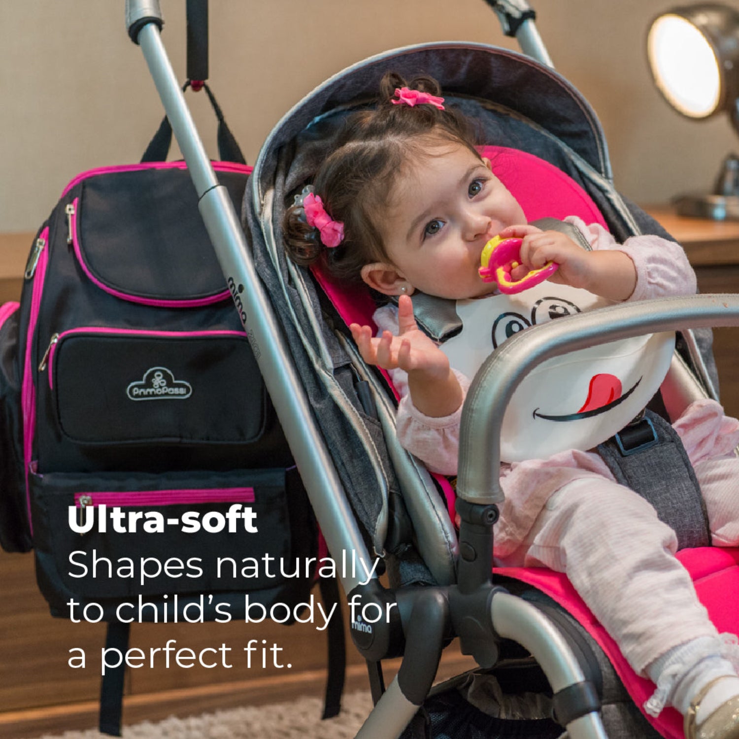 Toddler sitting comfortably in a stroller wearing a white Primo Passi silicone bib with a playful face design while chewing on a colorful teething toy. The ultra-soft silicone bib naturally conforms to the child's body for a comfortable fit, and the deep catch pocket helps keep clothes clean during snacks and meals. A Primo Passi diaper backpack is positioned beside the stroller, highlighting the bib as a practical travel essential in a cozy indoor setting.