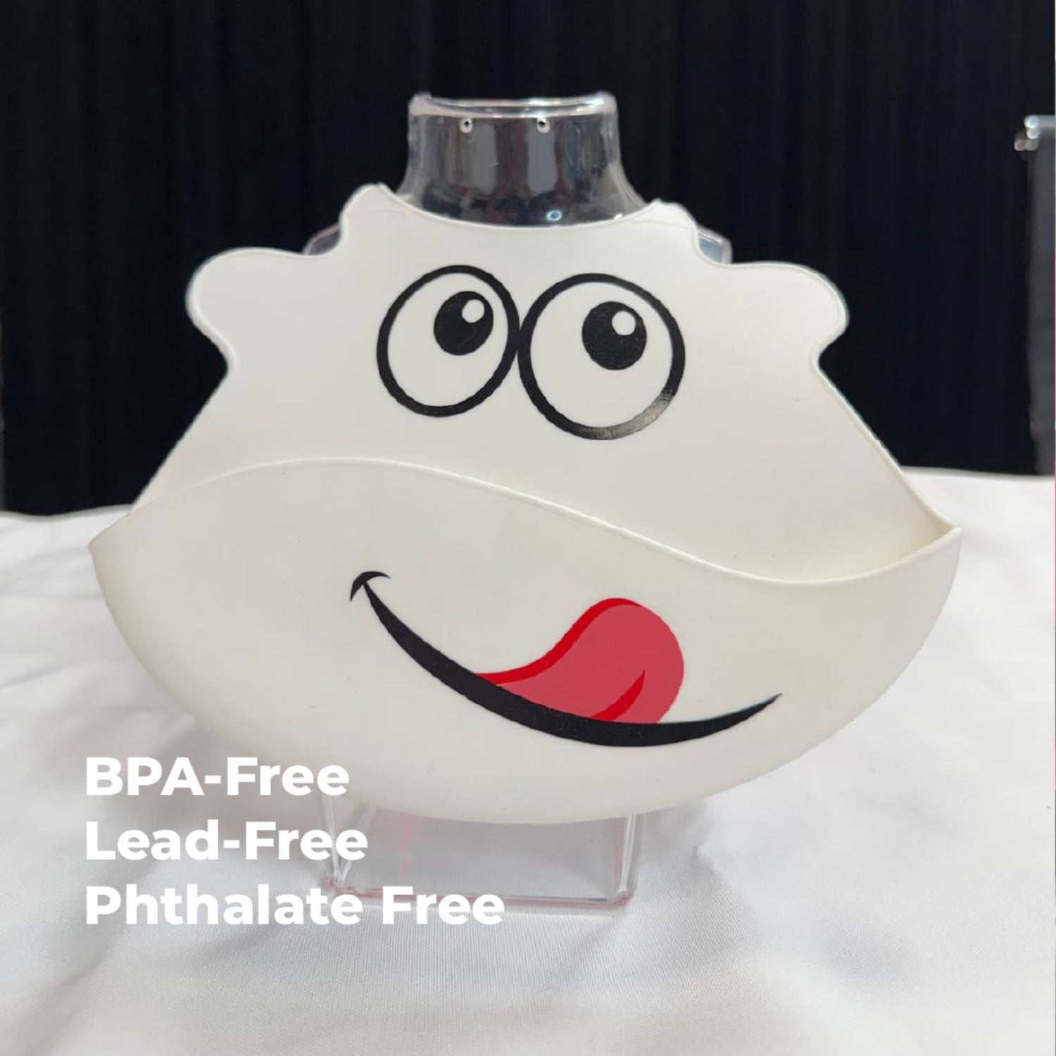 White Primo Passi silicone baby bib with a playful face design displayed on a clear stand over a white cloth surface with a dark background. The image highlights the bib's deep catch pocket and food-grade silicone construction, emphasizing that it is BPA-free, lead-free, and phthalate-free for safe, everyday feeding.