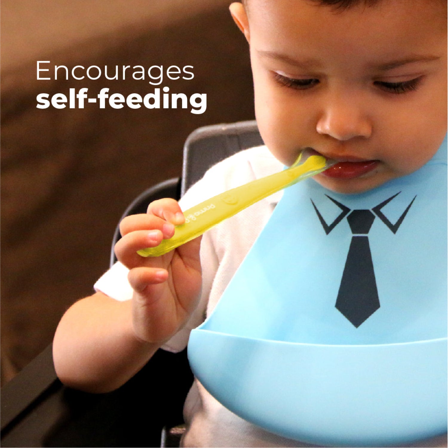 Toddler practicing self-feeding with a soft silicone baby spoon while wearing a silicone bib. The ultra-soft, food-grade silicone spoon is gentle on sensitive gums and designed to help babies build confidence and independence at mealtime.
