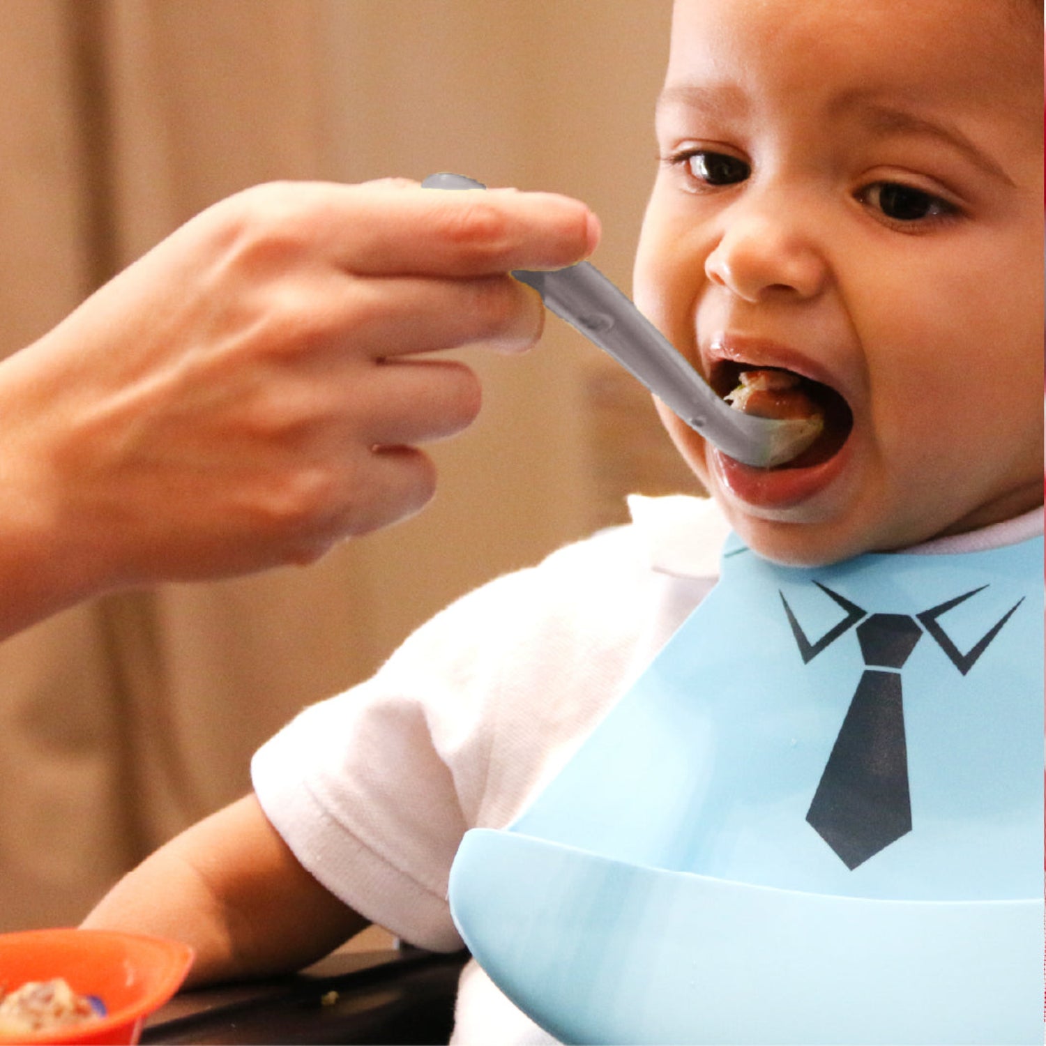 Parent feeding a toddler with a soft silicone baby spoon while the child wears a silicone bib. The ultra-soft, food-grade silicone spoon is gentle on sensitive gums and ideal for introducing solids and comfortable everyday feeding.