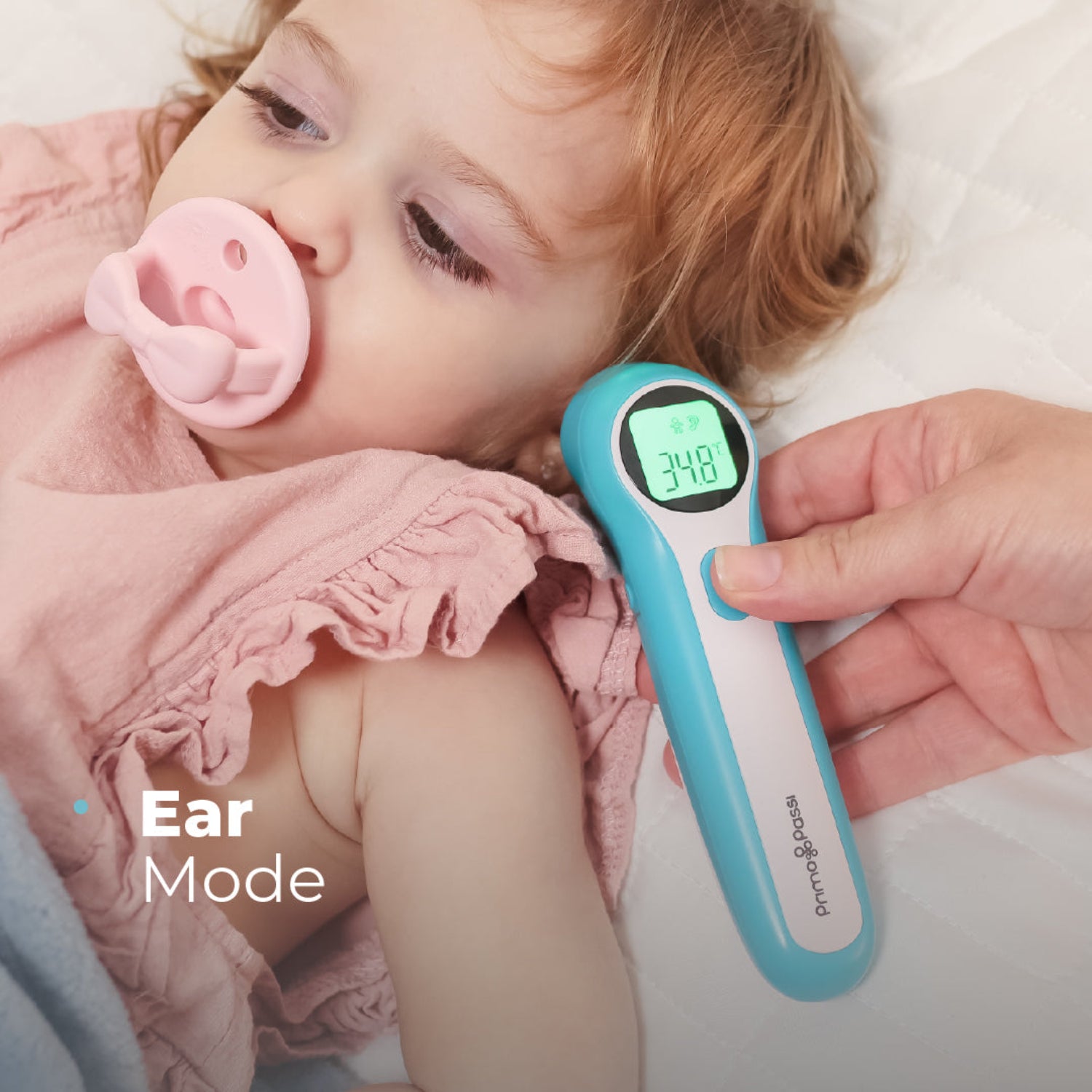A close-up lifestyle shot of a baby lying down with a pink pacifier, while an adult hand holds a blue and white digital ear thermometer. The thermometer displays a temperature of 34.8.