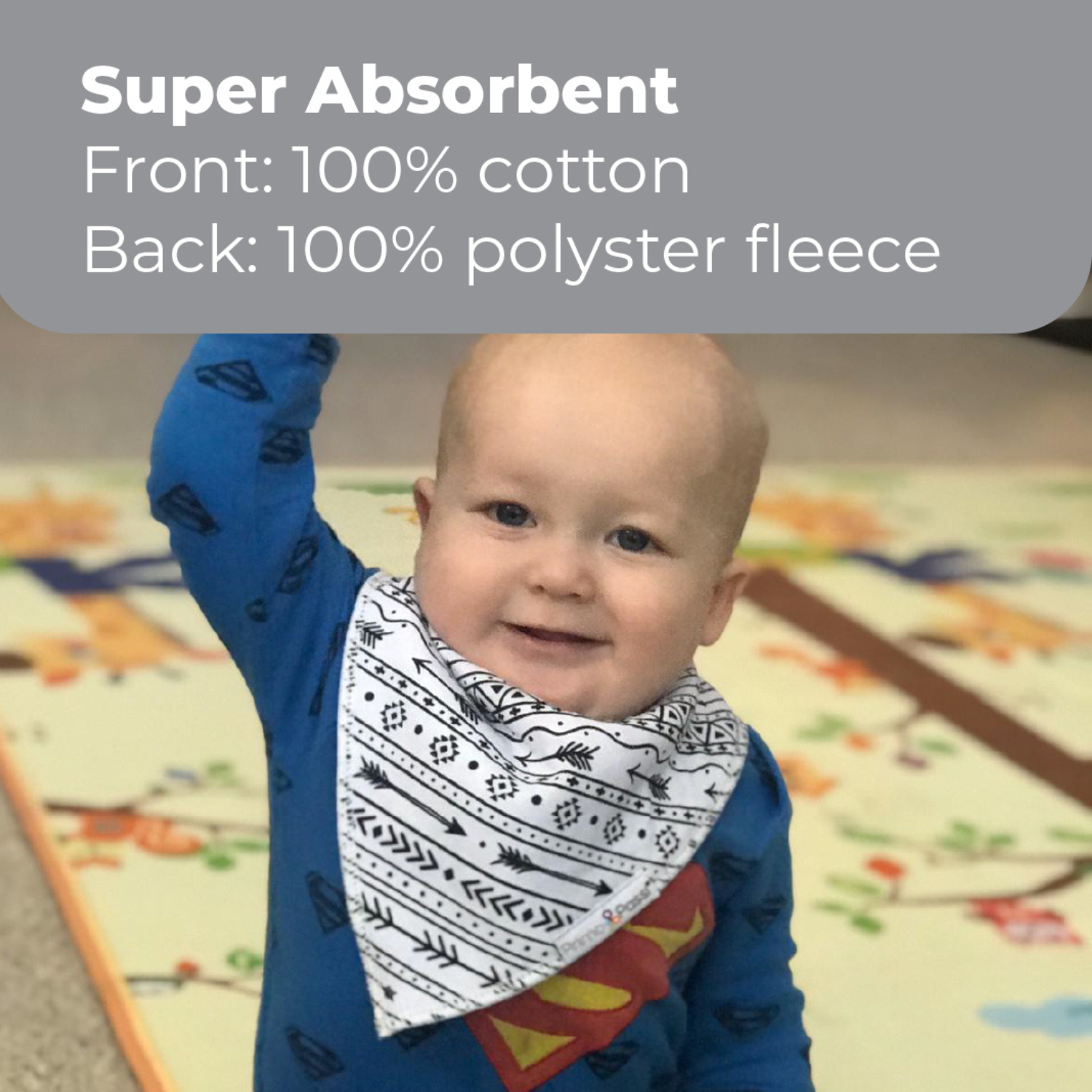 A smiling baby wearing a Primo Passi black-and-white tribal print bandana bib raises one arm while sitting on a colorful play mat. The bib features a super absorbent design with a 100% cotton front and 100% polyester fleece backing to help keep baby dry and comfortable throughout the day.