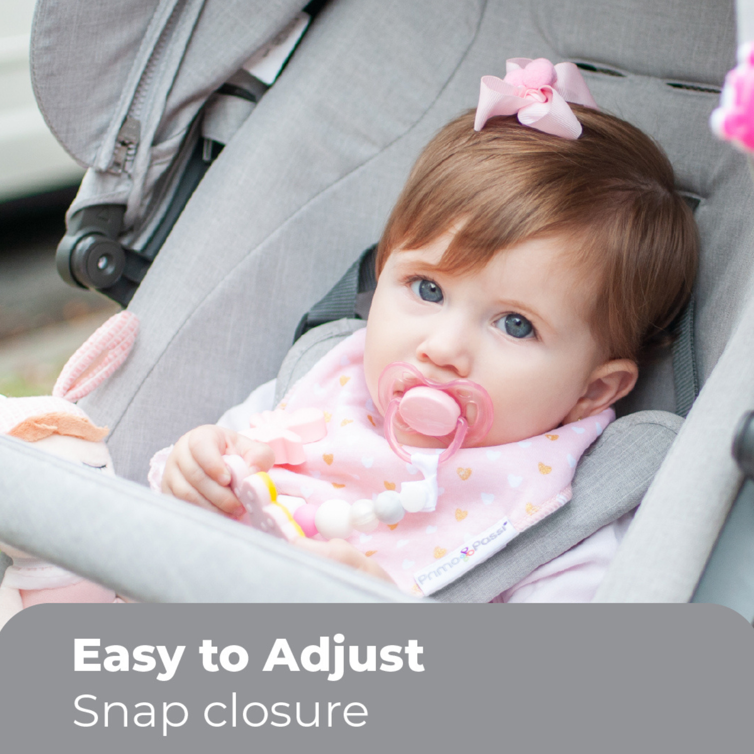 Baby wearing a Primo Passi pink bandana bib with a heart pattern while sitting comfortably in a stroller. The bib features an easy-to-adjust snap closure, an absorbent 100% cotton front, and a soft fleece backing to help keep clothing dry during teething, drooling, and everyday outings. Suitable for babies 0–9 months.