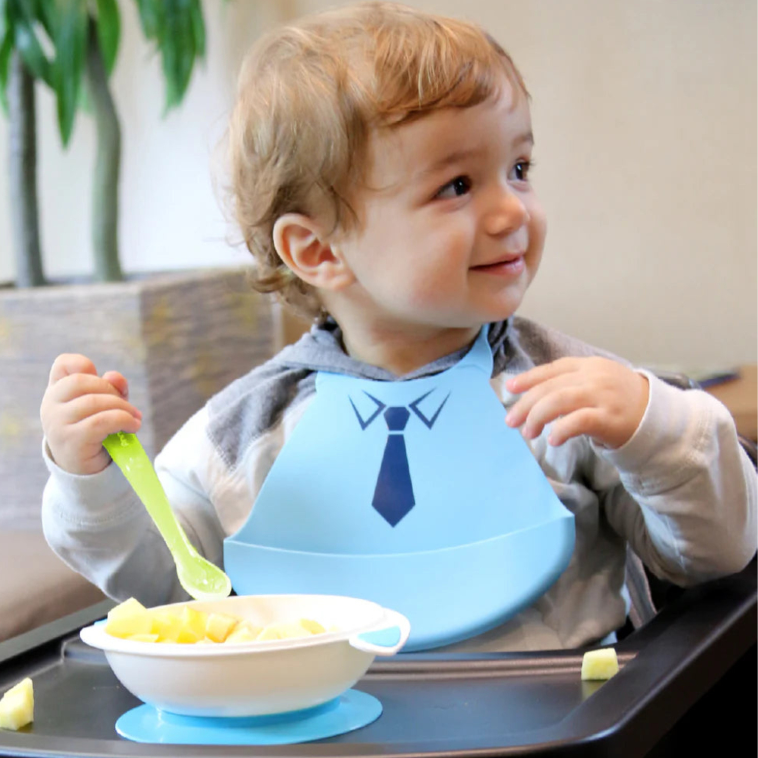 Child wearing a blue bib with a necktie design, eating from a silicone suction bowl with a green silicone spoon.