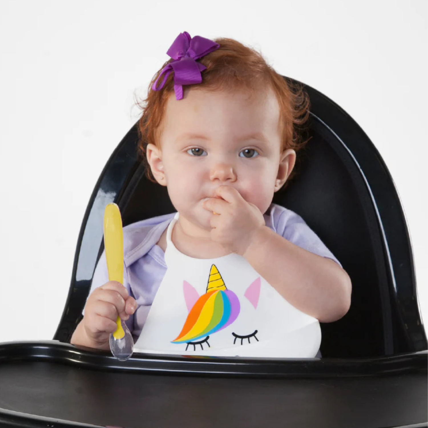 Child in a high chair with a rainbow unicorn bib, holding a yellow silicone spoon.