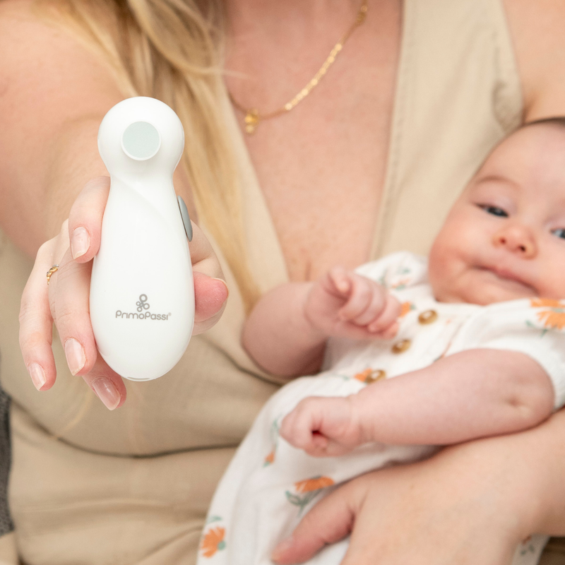 Close-up of a mother gently holding her baby's hand while using a white electric baby nail trimmer to safely file the baby's nails. The baby is cradled in her arms, creating a warm and comforting caregiving moment.