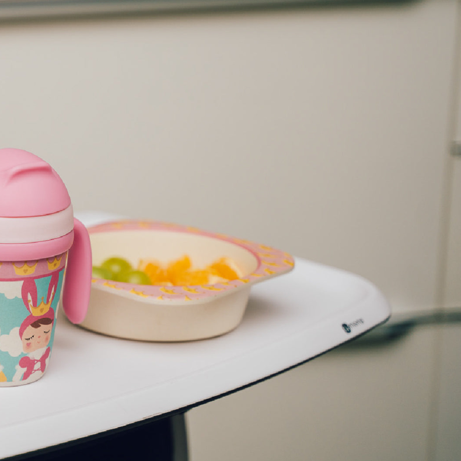 A pink toddler training cup and a matching bamboo fiber children's bowl with a princess-themed design sit on a high chair tray. The bowl is filled with fresh fruit, creating a coordinated, kid-friendly mealtime setup.