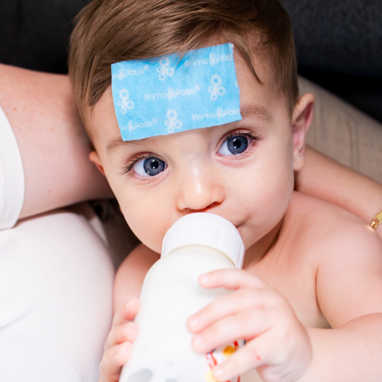 Close-up of a baby drinking from a bottle while wearing a cooling relief patch on the forehead. The cooling patch is designed to provide soothing comfort during fever or warm weather and stays securely in place while the baby rests.