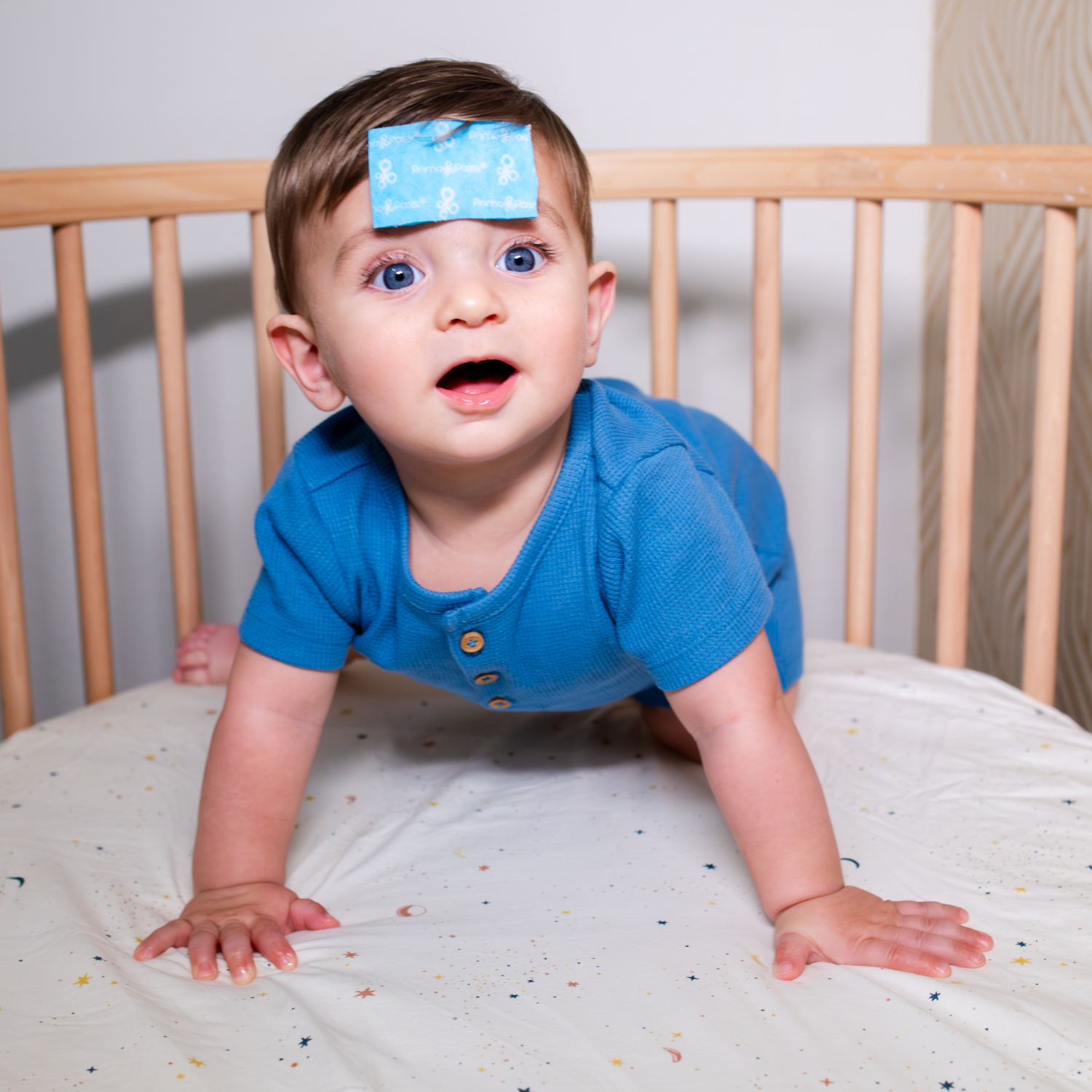 Baby crawling in a crib with a cooling relief patch on the forehead, designed to help provide soothing comfort during fever or warm weather. The child wears a blue romper and looks alert while the cooling patch stays securely in place.
