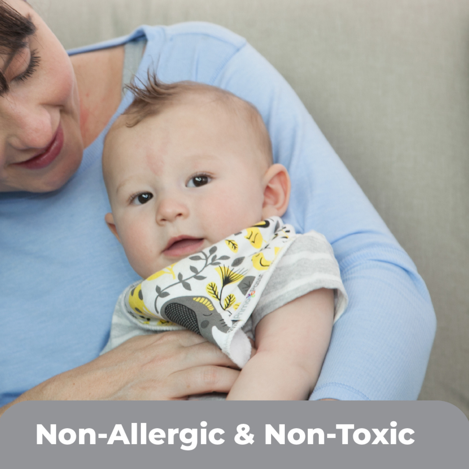 Baby wearing a Primo Passi bandana bib with a playful gray and yellow animal print while being held by a caregiver. The soft, non-allergenic and non-toxic bib helps protect clothing from drool and spills, making it ideal for everyday feeding and teething.